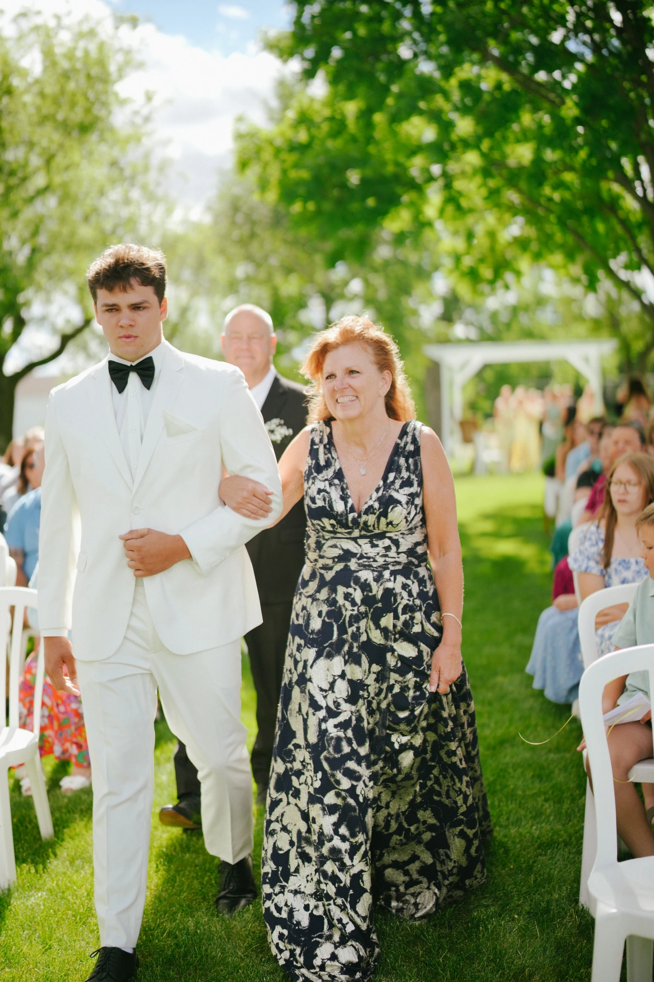 Teddy in his white suit walking with mother aisle at Amy and Teddy's North Dakota farm wedding by Megaset Weddings